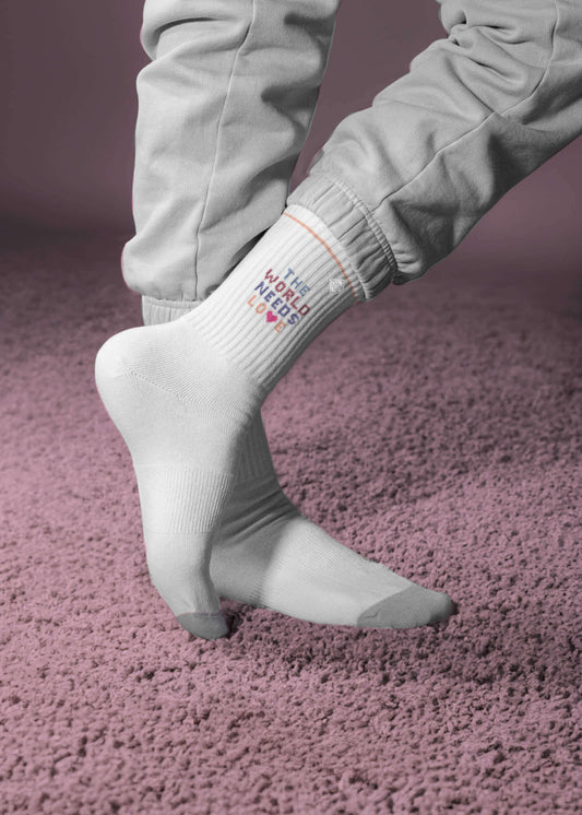 Ein Paar weiße J.Clay Tennissocken mit grauen Zehen und Absätzen, mit der Aufschrift "THE WORLD NEEDS LOVE" in bunten Blockbuchstaben und einem kleinen Herz unter dem Text. - Tennissocken