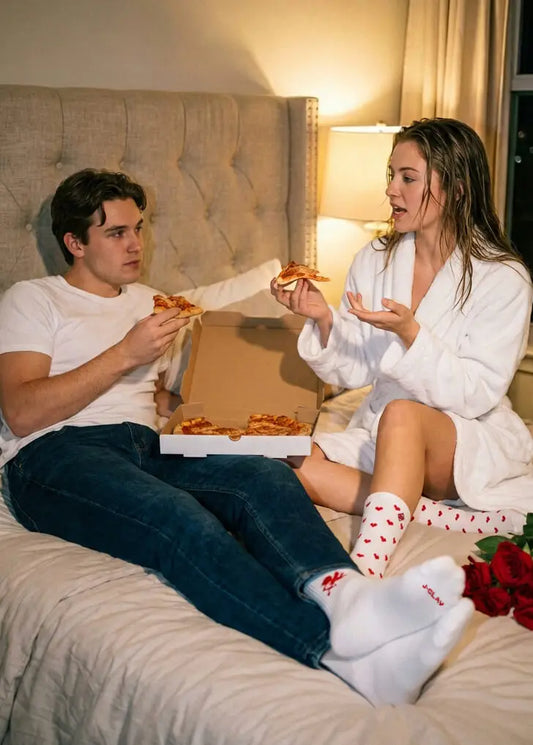 Ein Paar isst Pizza auf einem Bett; sie trägt einen Bademantel und J.Clay Red Hearts-Socken, er trägt Jeans.