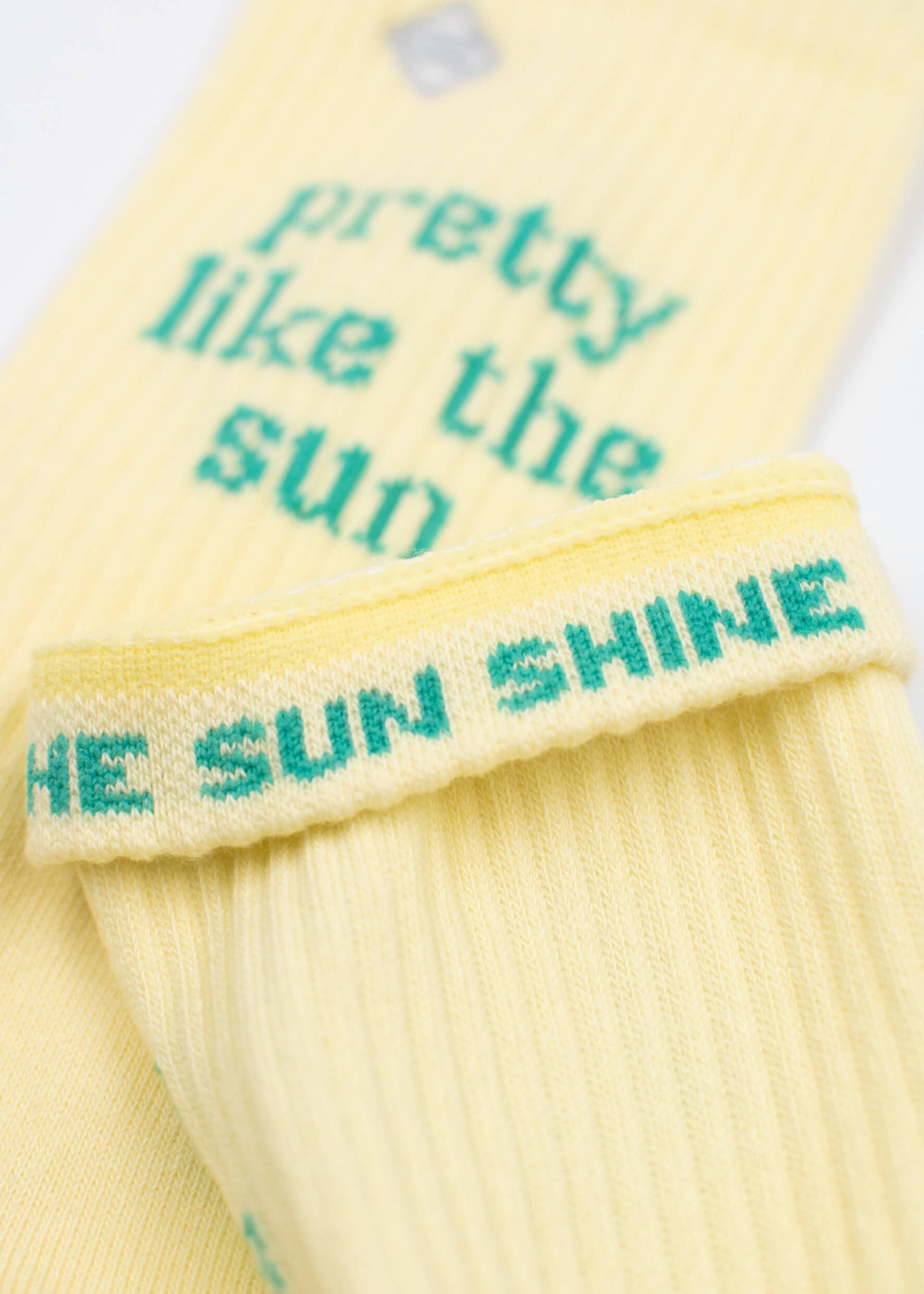 Nahaufnahme von gelben J.Clay-Tennissocken mit grünem Text - auf einer Socke steht "hübsch wie die Sonne", auf dem anderen Bündchen steht "THE SUN SHINE".
