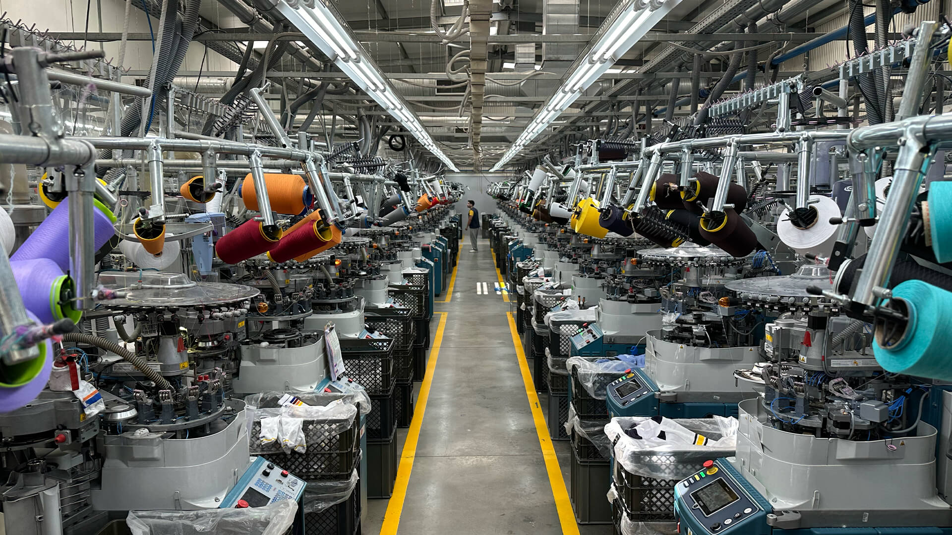 Eine Fabrik mit Reihen von Textilmaschinen und bunten Garnspulen, die eine symmetrische Industrieszene mit heller Deckenbeleuchtung und gelben Linien auf dem Boden bilden, die die Laufwege markieren.