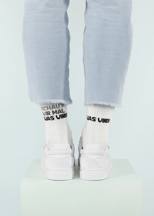 Eine Person, die eine hellblaue Cordhose von J.Clay, Tennissocken mit schwarzem Text und weiße Turnschuhe trägt, steht auf einem weißen Podest vor einem hellen Hintergrund; nur die Unterschenkel und Füße sind sichtbar.