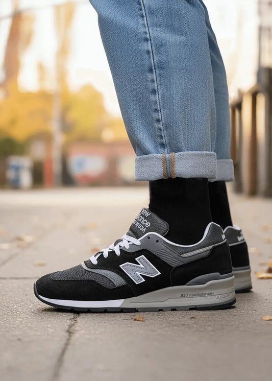 Nahaufnahme einer Person, die J.Clay Essential Black Socken, schwarze und graue New Balance 997 Turnschuhe und Blue Jeans mit Bündchen trägt und auf einem gepflasterten Bürgersteig vor einem unscharfen Hintergrund im Freien steht.