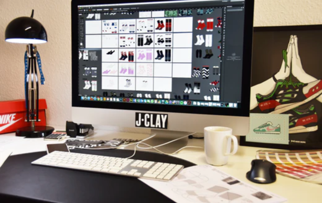 Ein Desktop-Computer zeigt ein Raster von Turnschuhdesigns in einem Arbeitsbereich mit Tastatur, Maus, Kaffeetasse, Schreibtischlampe, Schuhskizzen und gerahmten Kunstwerken von betenden Händen, die Turnschuhe halten.