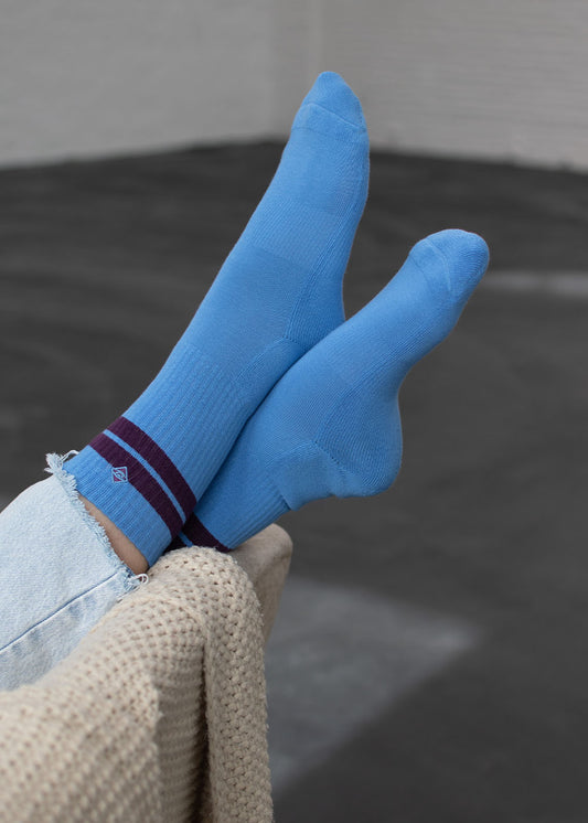 Die Füße ruhen auf einer beigen Decke, bekleidet mit J.Clay True Sky Grape Tennissocken.