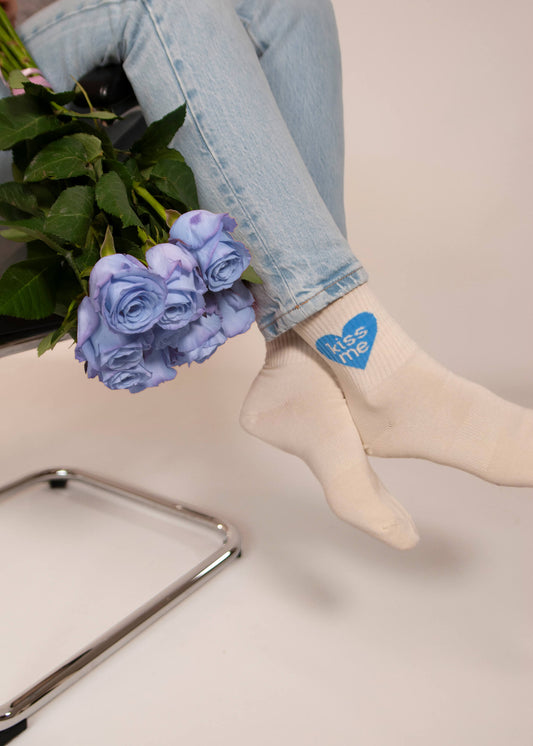 In hellblauen Jeans und cremefarbenen Tennissocken von J.Clay mit einem blauen "Kiss me"-Herz ruht jemand seine Füße auf einem Stuhl aus und hält einen Strauß hellvioletter Rosen in der Hand.