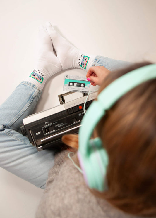 Eine Person mit mintgrünen Kopfhörern von J.Clay, hellblauen Jeans und weißen Tennissocken mit bunten Mustern sitzt im Schneidersitz und hält eine Kassette und einen alten Kassettenspieler auf dem Schoß.