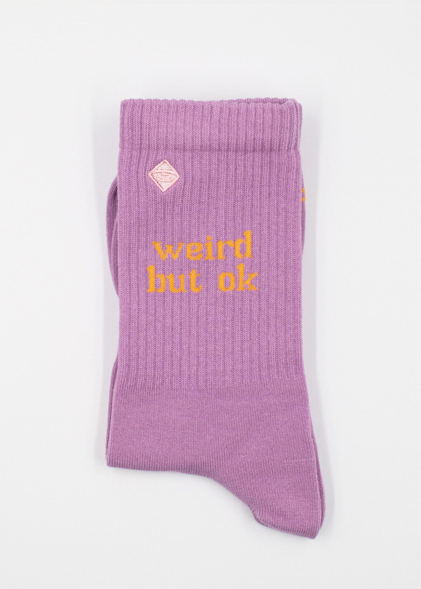 Ein Paar gefaltete lilafarbene J.Clay Tennissocken mit "weird but ok" in gelber Schrift auf der Seite und einem kleinen rosa rautenförmigen Logo über dem Text, dargestellt vor einem schlichten weißen Hintergrund.