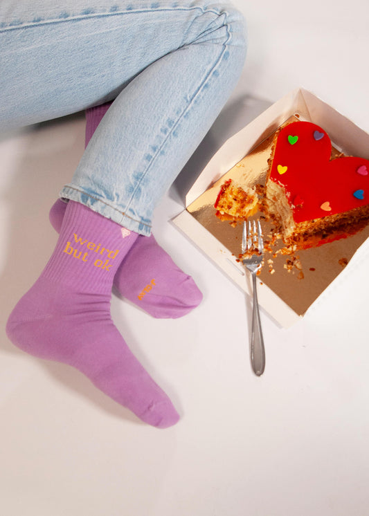 Eine Person in hellvioletten J.Clay-Tennissocken mit gelber Aufschrift und hellblauen Jeans sitzt neben einem teilweise aufgegessenen herzförmigen Kuchen, der mit bunten Herzen belegt ist, und stützt eine Gabel auf die Schachtel.