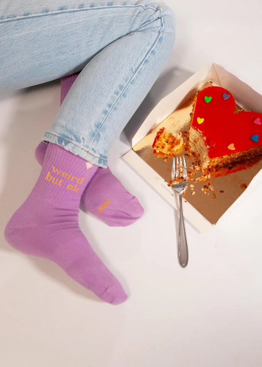 Eine Person in hellvioletten J.Clay-Tennissocken mit gelber Aufschrift und hellblauen Jeans sitzt neben einem teilweise aufgegessenen herzförmigen Kuchen, der mit bunten Herzen belegt ist, und stützt eine Gabel auf die Schachtel.