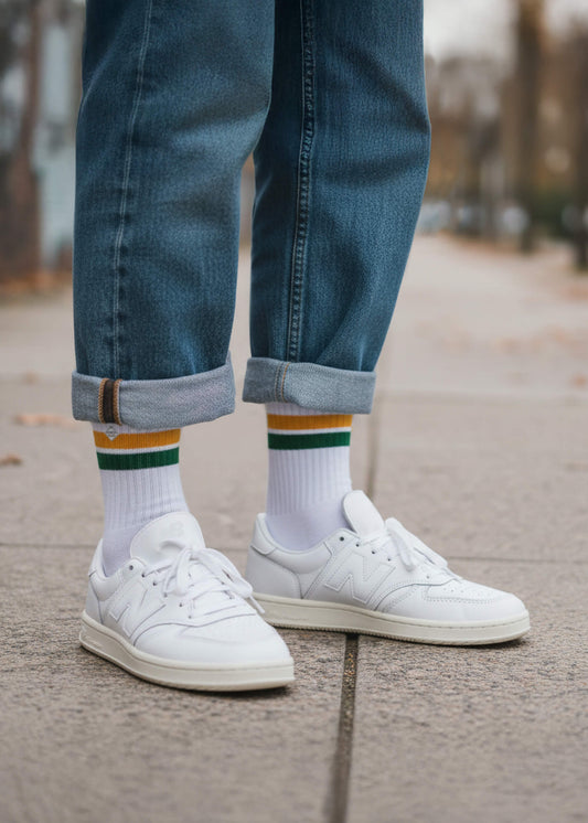 Eine Person mit Jeanshose und weißen New Balance Schuhen trägt die weißen Tennissocken mit orangen und grünen Streifen
