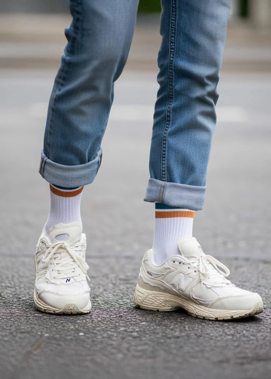 Eine Person mit Jeanshose und beigen New Balance Schuhen trägt die weißen Tennissocken mit petrolfarbenden und orangen Streifen