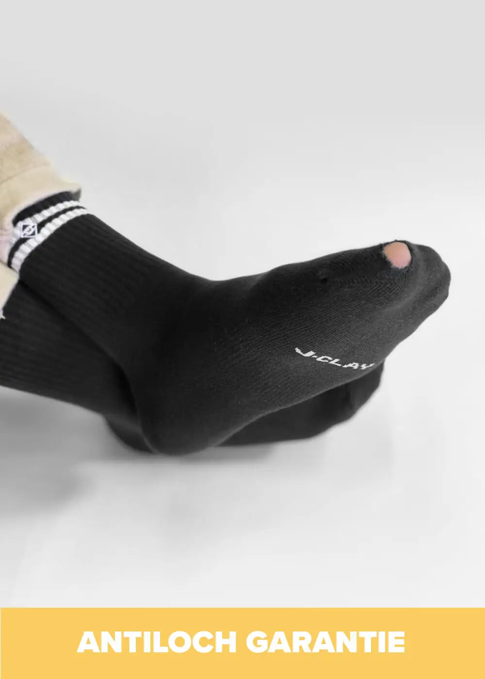 Eine Person trägt schwarze J.Clay-Tennissocken mit einem sichtbaren Loch an einem Zeh. Der Hintergrund ist einfarbig, und "ANTILOCH GARANTIE" erscheint in fetter weißer Schrift auf einem gelben Banner am unteren Rand.