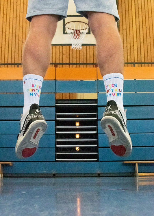 Eine Person springt in einer Turnhalle und trägt weiße J.Clay-Tennissocken mit der Aufschrift "Bitch Don't Kill My Vibe", graue Shorts und schwarz-weiße Turnschuhe. Im Hintergrund sind ein Basketballkorb und eine blaue Tribüne zu sehen.
