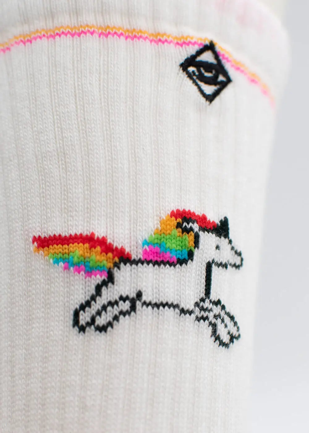 Nahaufnahme einer weißen J.Clay-Tennissocke mit einem kleinen schwarz umrandeten Einhorn mit Regenbogenmähne und -schwanz, rosa und gelben Streifen in der Nähe der Spitze und einem schwarzen gestickten Augensymbol oberhalb des Einhorns.