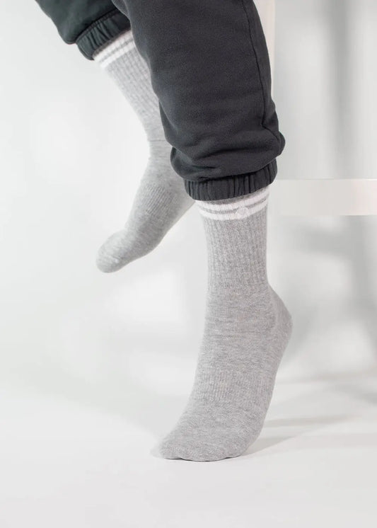 Eine Person trägt eine dunkelgraue Jogginghose von J.Clay und hellgraue Tennissocken mit weißen Streifen. Sie sitzt auf einer weißen Fläche, hat die Füße leicht gekreuzt und nur die Unterschenkel sind sichtbar.