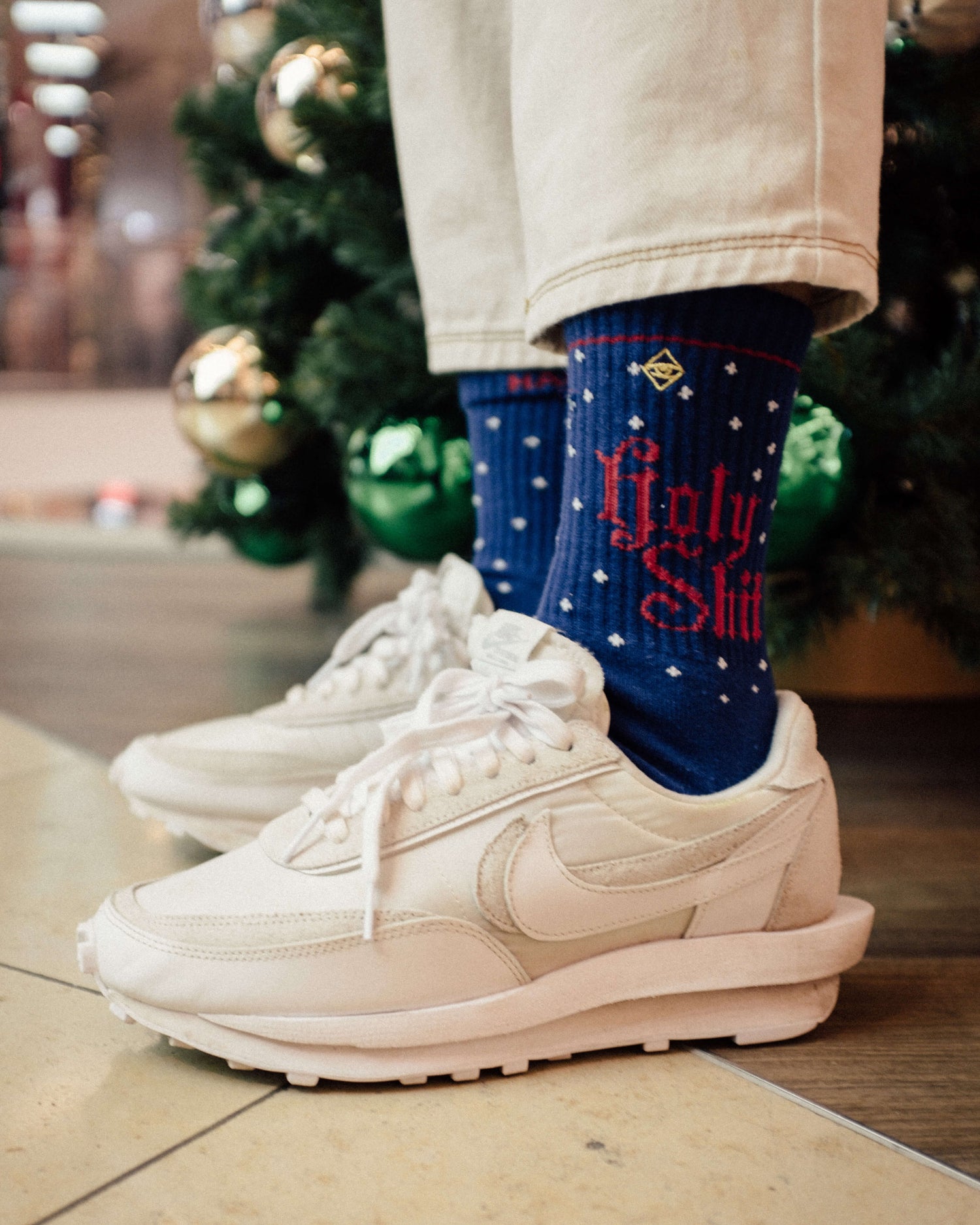 Eine Person in weißen Turnschuhen und cremefarbener Hose steht an einem Weihnachtsbaum; ihre blauen Socken sind mit Schneeflocken und der roten Aufschrift "Holy Shit" versehen. Im Hintergrund sind grüne Ornamente zu sehen.