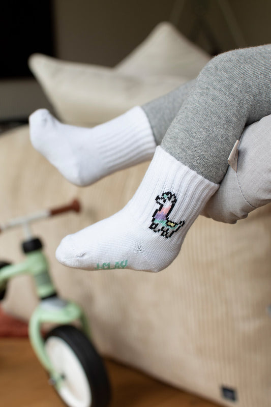 Ein Kind in grauer Hose und J.Clay Baby Socken mit einem kleinen Einhornmotiv sitzt auf einem hellen Kissen, im Hintergrund ein mintgrünes Dreirad.