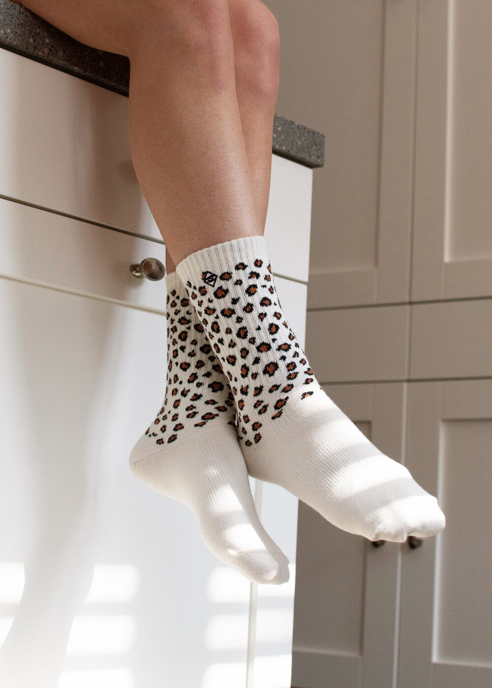 Eine Person sitzt auf einem Küchentisch und trägt weiße Tennissocken von J.Clay mit einem schwarz-braunen Leopardenmuster. Das Sonnenlicht wirft Schatten auf die weißen Schränke im Hintergrund.