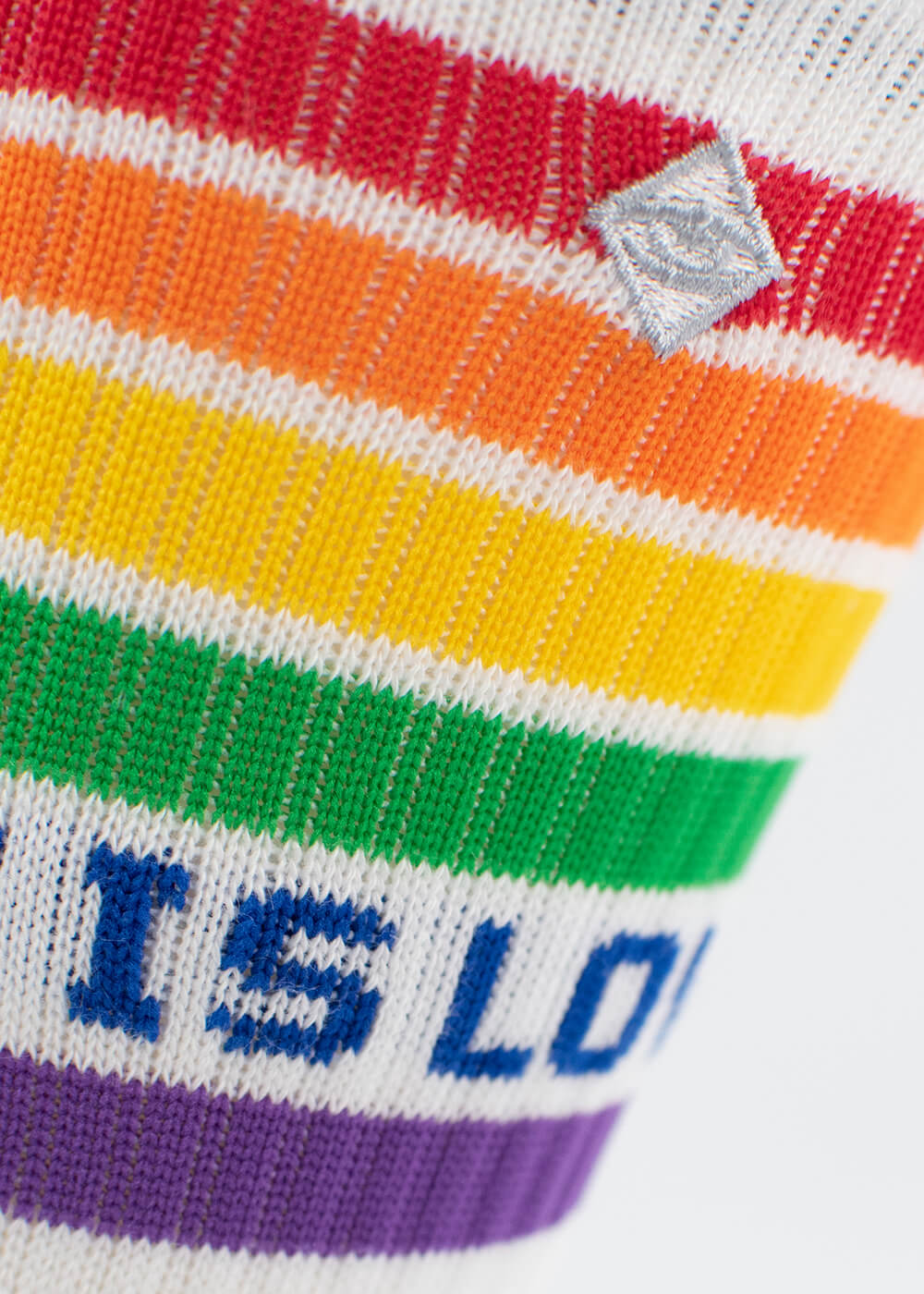 Nahaufnahme eines gestrickten Tennissockenstoffs von J.Clay mit horizontalen Regenbogenstreifen und teilweise blauem Text. "IS LO" ist sichtbar, zusammen mit einem kleinen rautenförmigen Aufnäher oberhalb der Streifen.