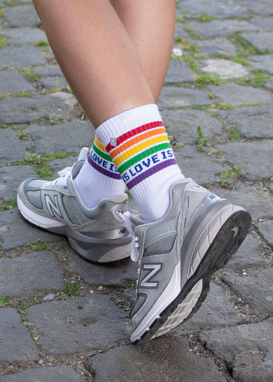 Eine Person geht in grauen New-Balance-Turnschuhen und J.Clay-Tennissocken - weiße Socken mit Regenbogenstreifen und der Aufschrift "LOVE IS LOVE IS" - über einen kopfsteingepflasterten Weg, zwischen dessen Steinen grünes Moos wächst.