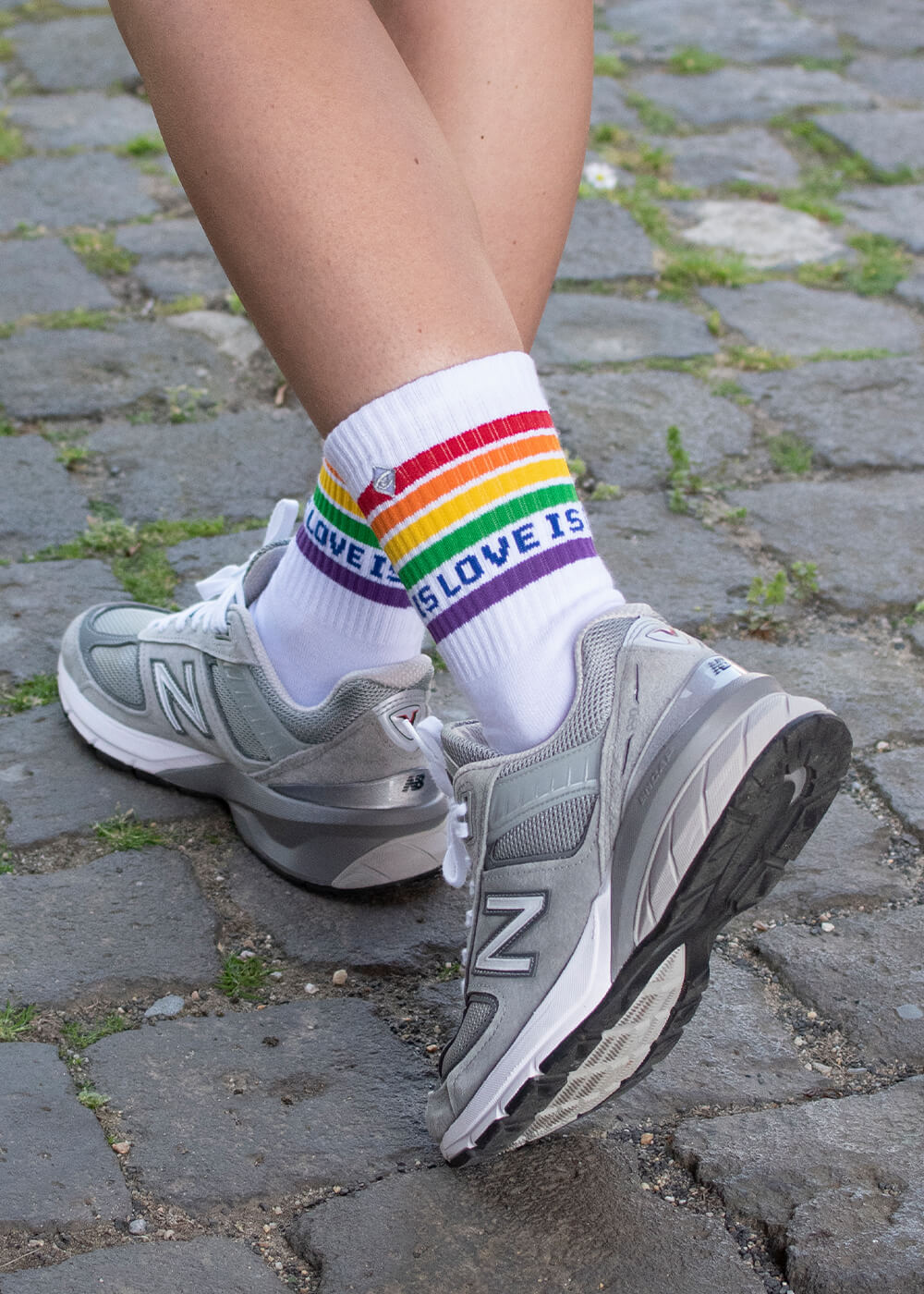 Eine Person geht in grauen New-Balance-Turnschuhen und J.Clay-Tennissocken - weiße Socken mit Regenbogenstreifen und der Aufschrift "LOVE IS LOVE IS" - über einen kopfsteingepflasterten Weg, zwischen dessen Steinen grünes Moos wächst.