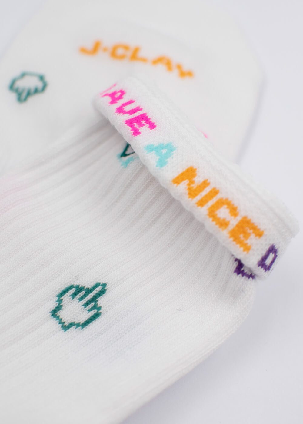 Nahaufnahme einer weißen gerippten J.Clay-Tennissocke mit dem bunten Text "HAVE A NICE DAY" auf dem Bündchen, kleinen grünen Cursor-Symbolen auf dem Körper und einem Teil des J.Clay-Markennamens in Orange.