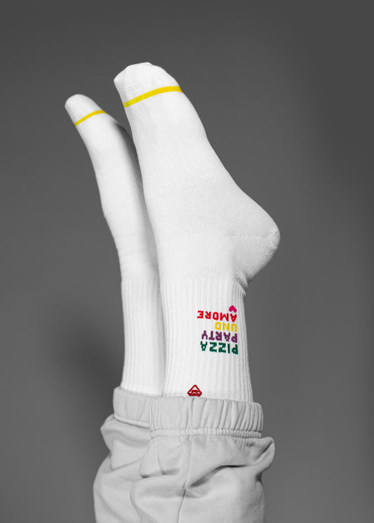 Zwei Füße in J.Clay Tennissocken - weiß mit gelben Zehenstreifen - zeigen den auf dem Kopf stehenden, bunten Text "PIZZA PARTY AND PANDEMIC". Die Trägerin trägt eine helle Hose auf grauem Grund.