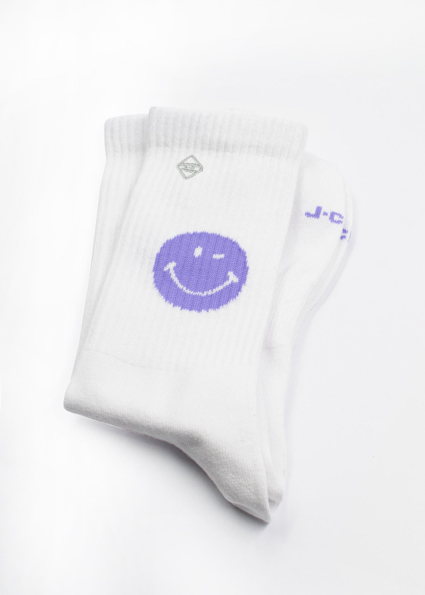 Ein Paar gefalteter weißer J.Clay Tennissocken mit einer großen lila Smiley-Grafik auf der Seite, dargestellt auf einem schlichten weißen Hintergrund.