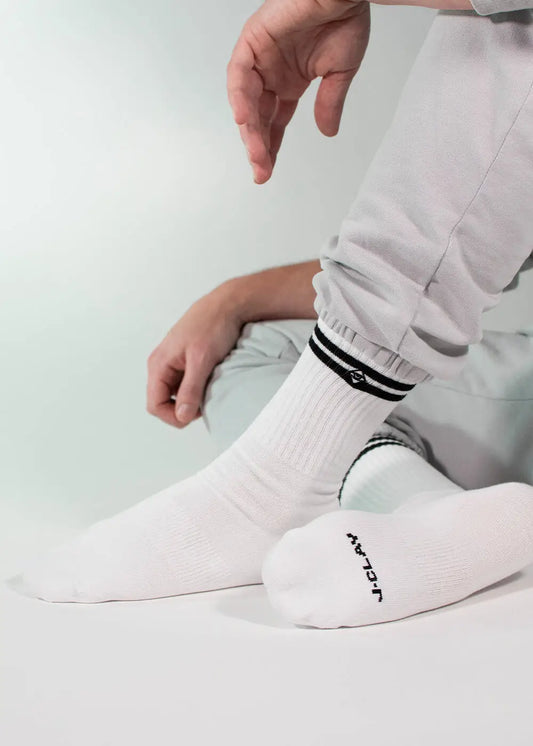 Eine Person in hellgrauer Hose sitzt mit einem angewinkelten Knie und trägt weiße J.Clay-Tennissocken mit schwarzen Streifen, einem kleinen Logo und "JCLAY" an der Sohle. Der Hintergrund ist einfarbig und hell gefärbt.