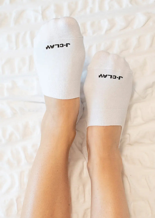 Zwei Füße in weißen J.Clay Unsichtbare Füßlinge mit schwarzem "LATER"-Schriftzug (auf der Rückseite) ruhen auf einer strukturierten weißen Decke. Diese rutschfesten Socken bieten zusätzlichen Komfort und Halt für den Alltag.