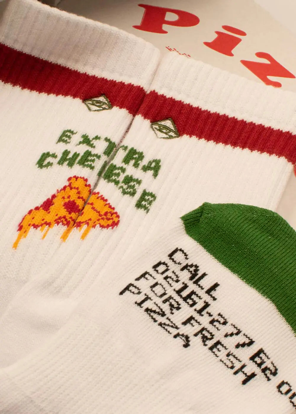 Nahaufnahme von zwei Paar J.Clay Tennissocken: ein Paar hat einen roten Streifen, Rauten, den Text "EXTRA CHEESE" und eine Pizzascheibe; das andere hat eine grüne Spitze und Ferse mit "CALL 025-277 FOR FRESH PIZZA".