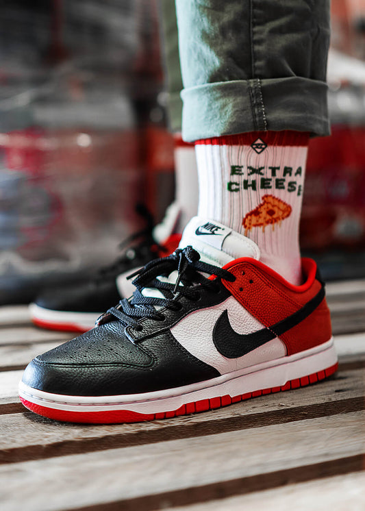 Eine Person, die schwarze, weiße und rote Nike-Turnschuhe und J.Clay-Tennissocken mit einer Pizzagrafik und dem Text EXTRA CHEESE trägt, steht auf einem Holzboden.