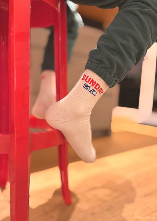 Eine Person in dunkelgrüner Hose sitzt auf einem leuchtend roten Hocker, die Füße ruhen auf dem Geländer, und trägt J.Clay Tennissocken mit SUNDAY CLUB in Rot und Blau. Der sanft verschwommene Hintergrund verleiht dem Bild eine sanfte Note.