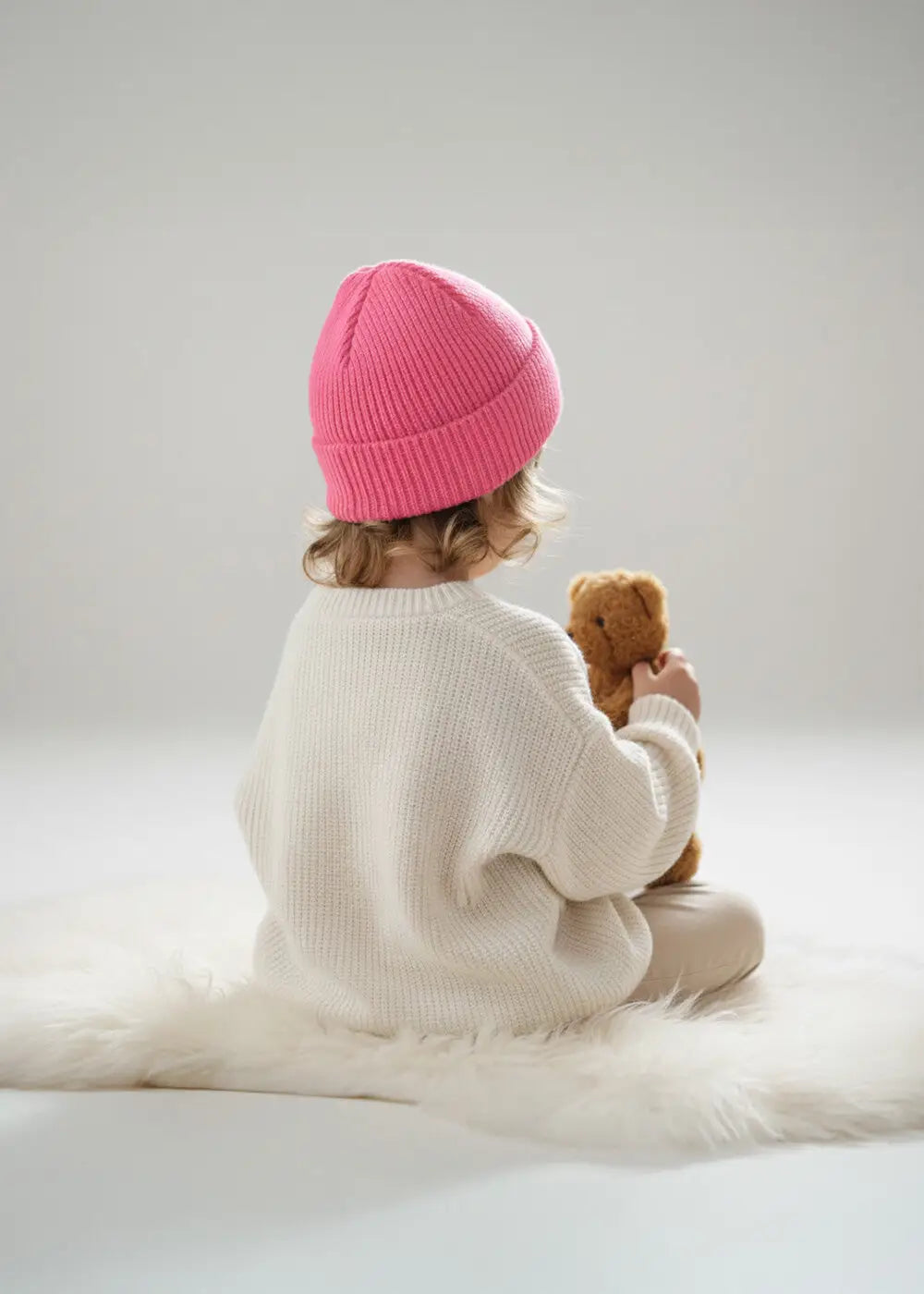 Ein kleines Kind mit einer rosa Strickmütze und einem weißen Pullover sitzt auf einem weichen weißen Teppich und hält einen kleinen braunen Teddybären. Der Hintergrund ist neutral und sanft beleuchtet.