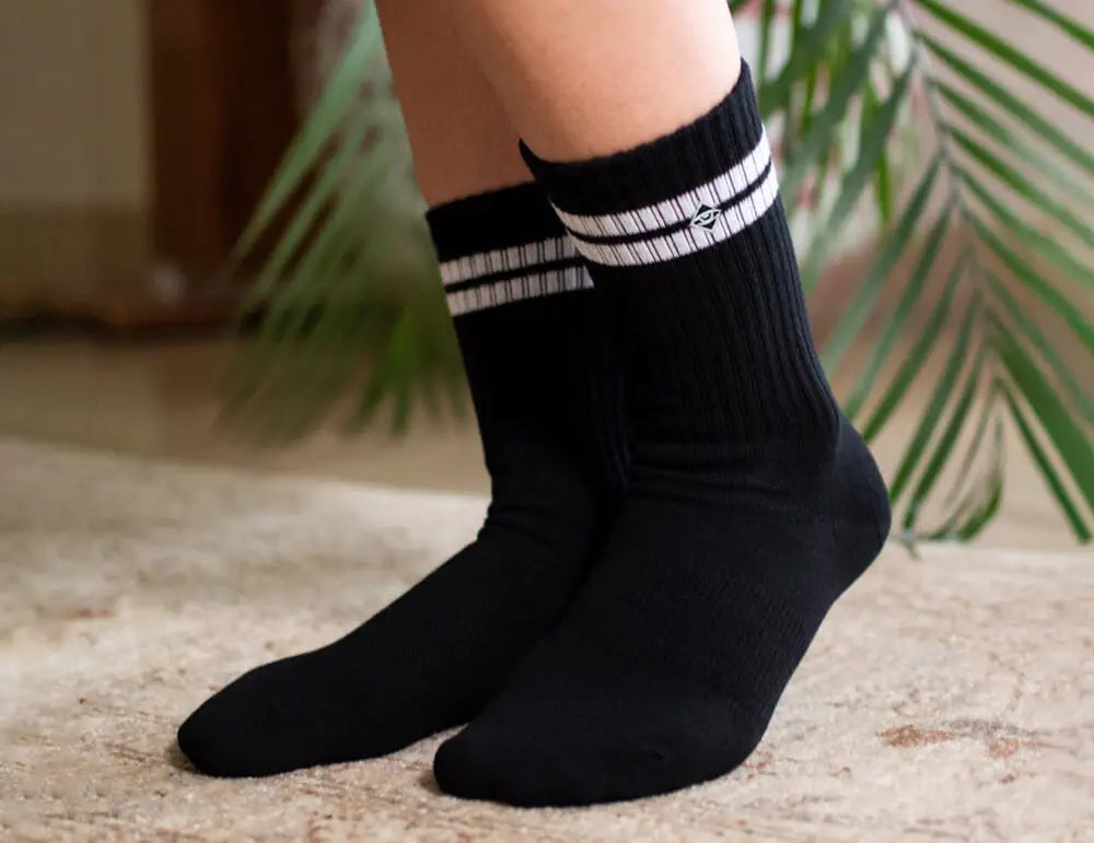 Basic Tennissocken für Damen - Nahaufnahme der Füße einer Person, die schwarze Socken mit weißen Streifen an der Spitze trägt und auf einem Teppichboden steht, mit grünen Pflanzenblättern im Hintergrund.