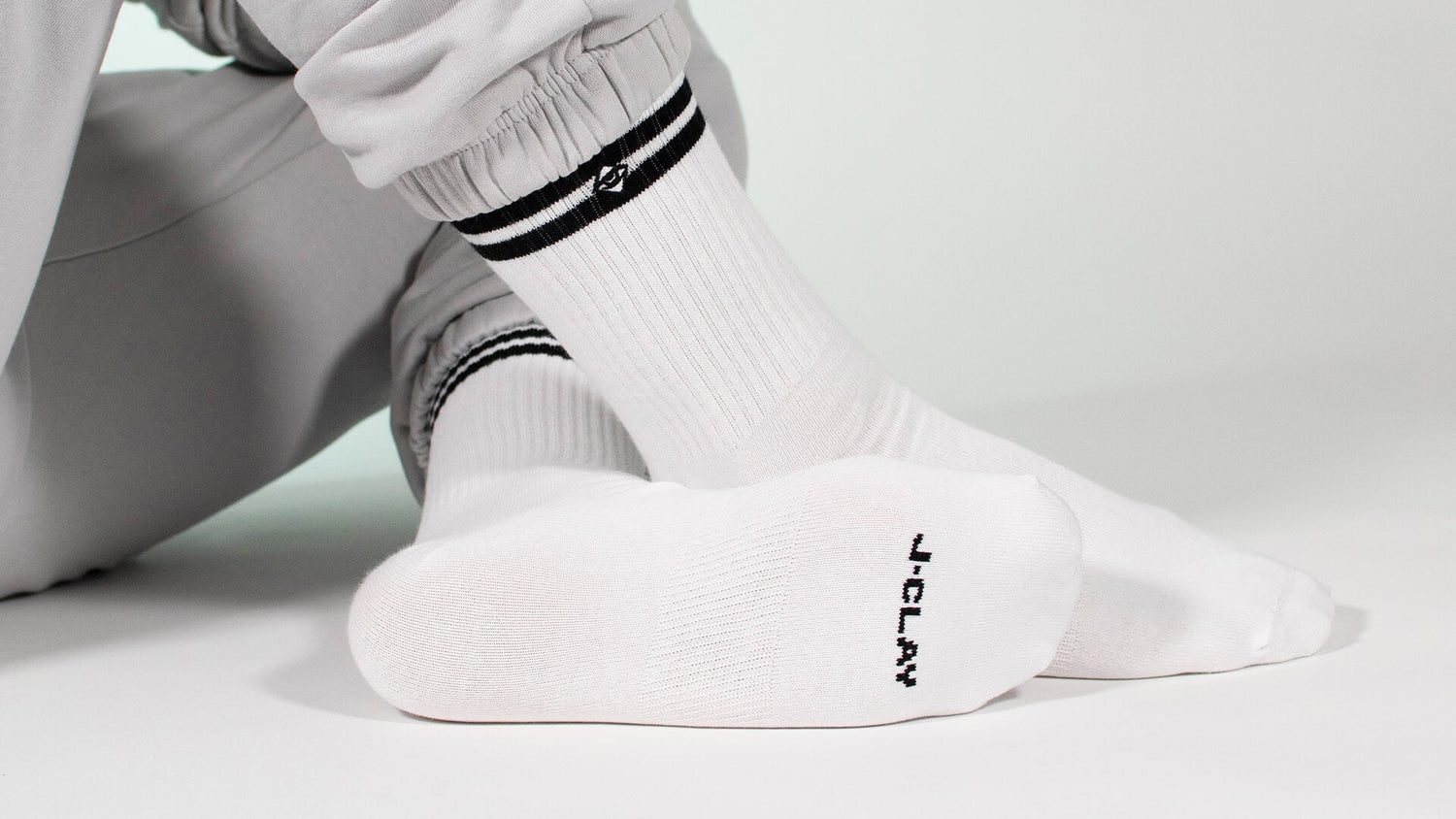 Weiße Tennissocken für Damen & Herren - Person, die weiße Socken mit schwarzen Streifen und Schriftzug an der Sohle trägt, mit gekreuzten Beinen auf dem Boden sitzt und mit einer hellgrauen Hose bekleidet ist.