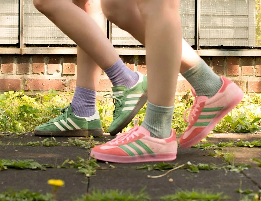 Zwei Personen gehen auf einem Weg, sie tragen pastellfarbene Turnschuhe - ein Paar grün, das andere rosa - und dazu hellviolette Socken. Zwischen Rissen im Pflaster wachsen Gras und Pflanzen, im Hintergrund ist eine Backsteinmauer zu sehen.