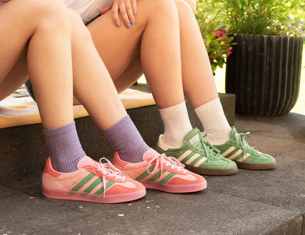 Zwei Personen, die auf einer Stufe im Freien sitzen und jeweils pastellfarbene Turnschuhe tragen - ein Paar in Rosa mit grünen Streifen und lila Socken, das andere in Grün mit weißen Streifen und weißen Socken. Im Hintergrund sind Pflanzen zu sehen.