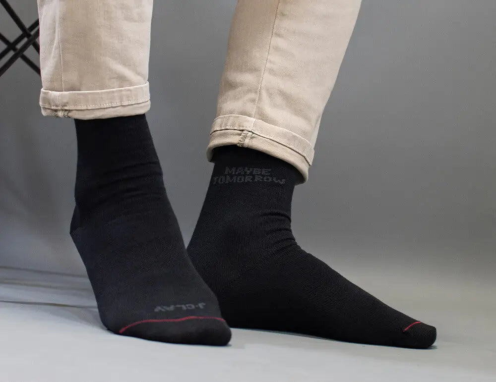 Business Socken für Herren - Person in beiger Hose und schwarzen Socken mit der Aufschrift MAYBE TOMORROW, die auf einer grauen Fläche sitzt. Nur die Unterschenkel und Füße sind sichtbar.