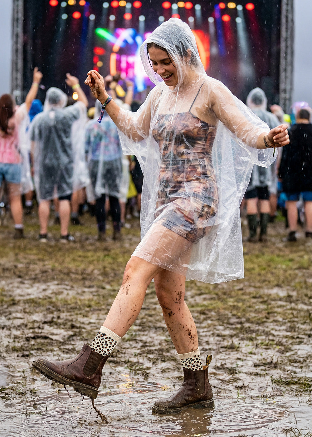 Eine Frau in einem Poncho und Stiefeln tanzt fröhlich auf einer schlammigen Wiese bei einem Musikfestival im Freien, mit bunten Bühnenlichtern und anderen Menschen im Hintergrund bei regnerischem Wetter.