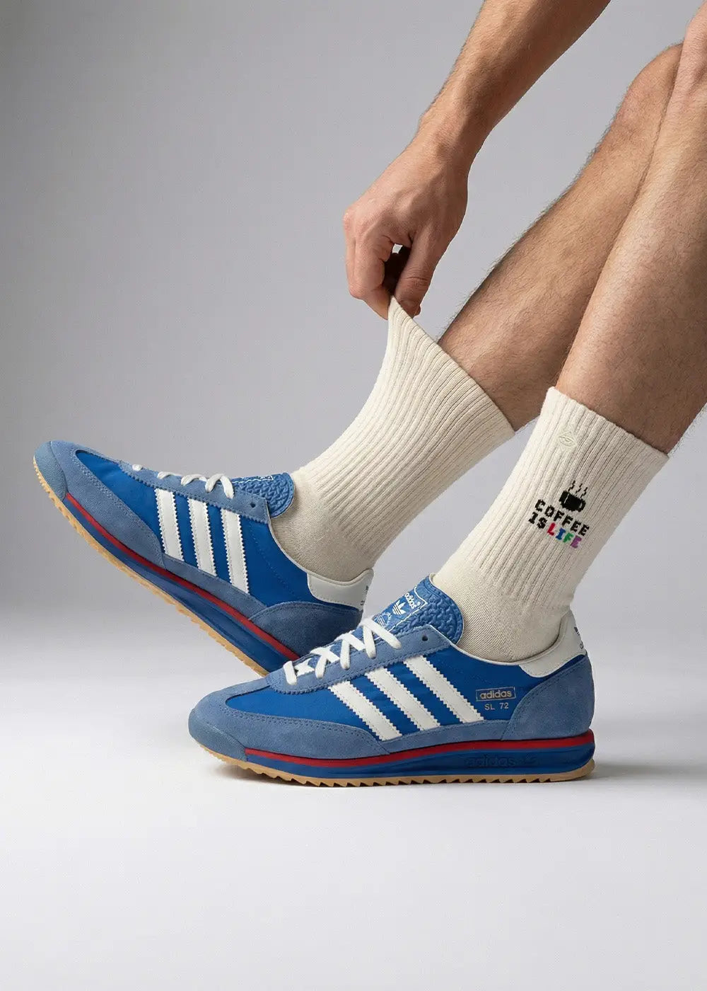 Eine Person, die weiße Socken mit der Aufschrift COFFEE & SOUL und blaue und weiße Adidas-Turnschuhe trägt, zieht eine Socke hoch, während sie vor einem schlichten Hintergrund sitzt.