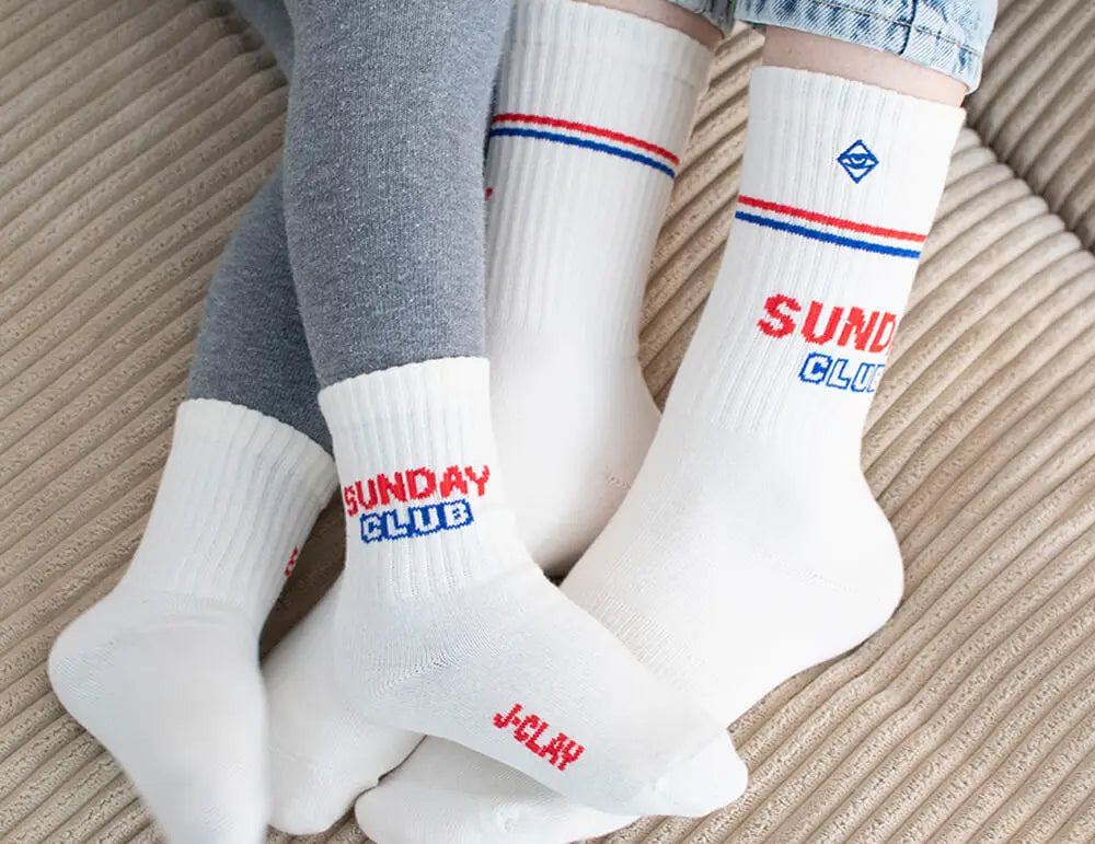 Zwei Personen, die zusammenpassende weiße Socken mit roten und blauen Streifen und dem Schriftzug SUNDAY CLUB an den Seiten tragen, sitzen zusammen auf einer beigefarbenen, strukturierten Fläche.