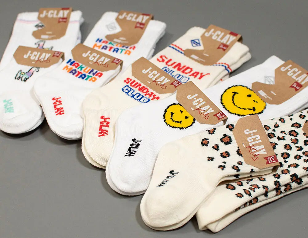 Fünf Paar weiße Socken mit farbenfrohen Motiven und Mustern, darunter Smiley-Gesichter, Tierdrucke und verspielte Texte, auf einer grauen Oberfläche mit J-CLAY Markenanhängern.
