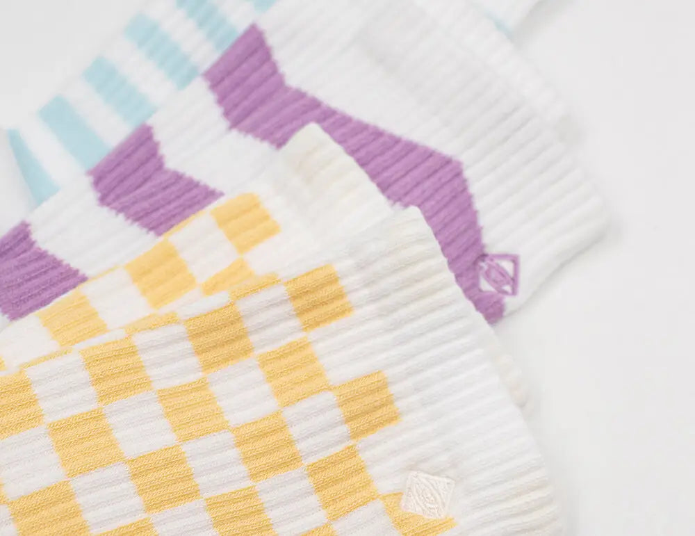 Eine Nahaufnahme von drei Paar gerippten Socken mit geometrischen Mustern aus blauen Streifen, violetten Chevrons und gelben Schachbrettern auf weißem Grund, die übereinander gelegt sind.