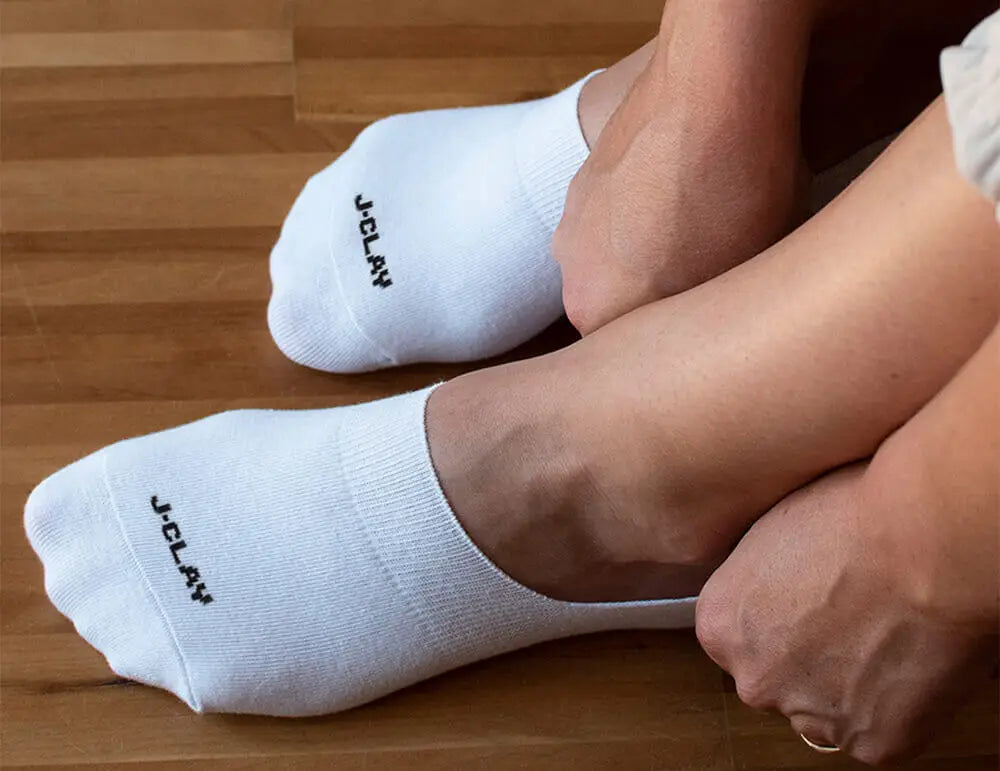 Füßlinge für Damen - Eine Person, die weiße Socken mit der Aufschrift J-CLAY trägt, auf einem Holzboden sitzt und einen Knöchel mit der Hand festhält.