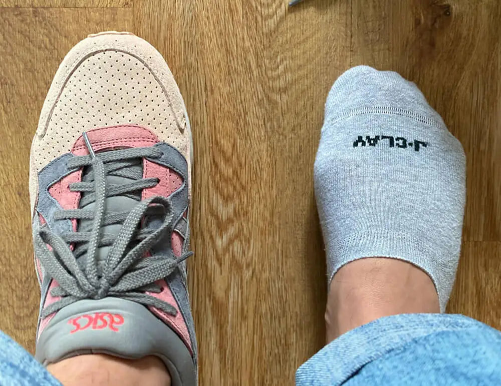 Eine Person in blauen Jeans hat einen grauen und rosa ASICS-Sneaker an einem Fuß und eine graue Socke mit schwarzer Schrift am anderen Fuß und steht auf einem Holzboden.