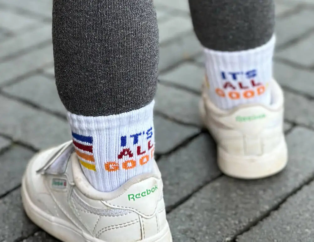 Eine Person in grauen Leggings, weißen Reebok-Turnschuhen und weißen Socken mit bunten Streifen und dem Text "IT'S ALL GOOD" steht auf einem grauen Backsteinpflaster.