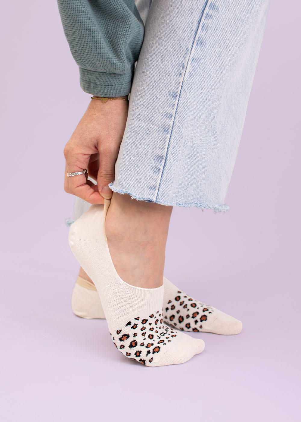 Kurze Socken - Eine Person in einer hellen, ausgefransten Jeans passt eine weiße Socke mit einem Leopardenmuster an den Zehen vor einem hellvioletten Hintergrund an.