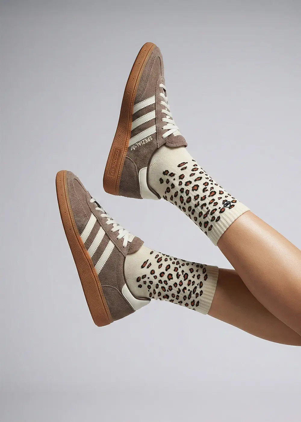 Eine Person, die cremefarbene Socken mit einem braunen Leopardenmuster und braune Adidas-Sneakers aus Wildleder mit weißen Streifen und Gummisohlen trägt, hebt die Beine vor einem neutralen Hintergrund.