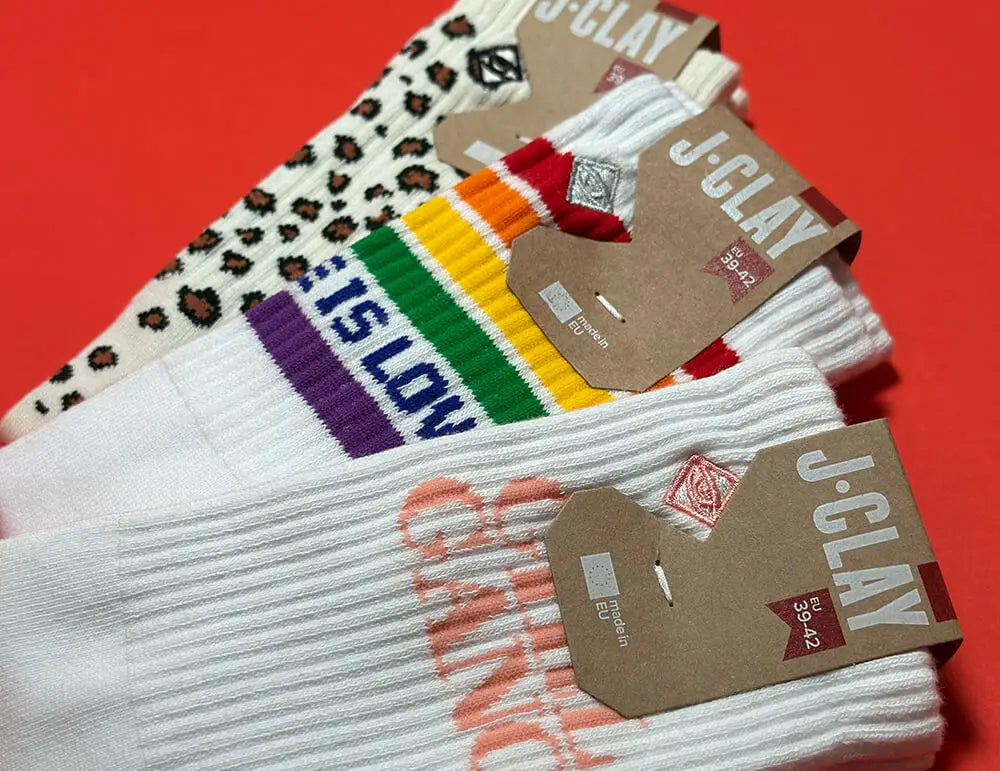 Socken Packs für Damen: Drei Paar JCLAY-Socken auf rotem Untergrund: eines mit Leopardenmuster, eines mit Regenbogenstreifen und "SLOW" und ein weißes Paar mit orangefarbenem Text, alle mit Pappanhänger.